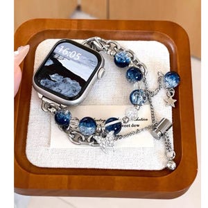 May include: Silver Apple Watch with a chain-style band featuring blue glass beads and star charms. The watch face displays the time as 16:05. The band includes the text "sweet dew".