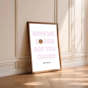 Op de afbeelding: Een ingelijste print met de tekst "GAVE ME COOKIE GOT YOU COOKIE" in roze, met een koekjesafbeelding. De naam van de kunstenaar, Nick Miller, staat onderaan. De lijst is bruin en de achtergrond is wit.