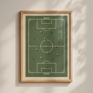 May include: A framed wall art print featuring an overhead view of a green soccer field with white boundary lines and markings. Small figures of players are scattered across the pitch in a top-down perspective, enclosed in a light wood frame.