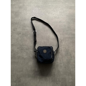 Op de afbeelding: Een marineblauwe crossbodytas met een zwarte verstelbare riem. De tas heeft een vierkante vorm met een hoofdvak met ritssluiting en een klein cirkelvormig logo aan de voorkant. De tas is gemaakt van een duurzaam, waterbestendig materiaal.