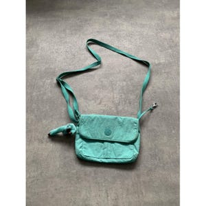 Peut inclure: Sac bandoulière turquoise avec fermeture à rabat et bandoulière réglable. Le sac présente un petit logo rond sur le devant et un porte-clés singe en peluche assorti. Le sac est en nylon froissé.