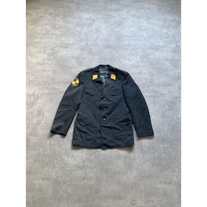 May include: A dark navy blue military jacket with gold shoulder patches and collar accents. The jacket has a button-up front, two chest pockets, and two lower pockets. The jacket is laid flat on a gray surface.