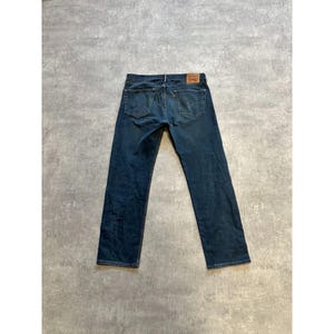 Pode incluir: Jeans de ganga de lavagem escura com um design clássico de cinco bolsos. Os jeans apresentam um remendo de couro castanho nas costas e uma etiqueta vermelha. Os jeans estão colocados sobre uma superfície cinzenta.