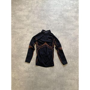 Puede incluir: Camiseta deportiva negra de manga larga con cuello alto. Presenta detalles marrones en los hombros y costuras naranjas. El nombre de la marca está impreso en el cuello.