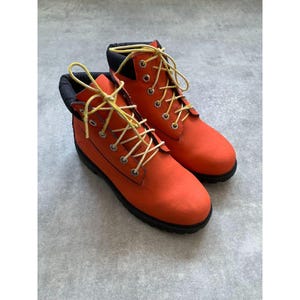 Puede incluir: Un par de botines de color naranja brillante con cuello acolchado negro, ojales metálicos plateados y cordones amarillos. Las botas tienen un diseño de punta redondeada y una suela de goma negra gruesa, ofreciendo una construcción robusta.
