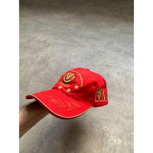 May include: A bright red baseball cap featuring gold embroidery on the front, including the text "WORLD CHAMPION" and "2002" with star accents. The brim displays a gold laurel leaf design, and a rectangular patch with a logo is on the side.