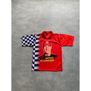 Pode incluir: Polo vermelho e preto de manga curta com um retrato de Michael Schumacher. A camisa tem um padrão xadrez na manga e no ombro esquerdos. O texto "MICHAEL SCHUMACHER" é impresso em amarelo.