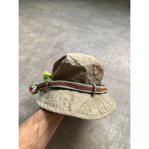 Può includere: Un cappello a secchiello beige con una toppa con logo in pelle marrone sul davanti e una fascia decorativa intrecciata attorno alla corona. La fascia presenta un motivo geometrico ripetuto in rosso, verde e bianco, rifinito con nappe sfrangiate.