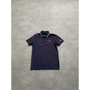 May include: A dark navy blue polo shirt with a white collar and a small white logo on the chest. The shirt has short sleeves and a Nike swoosh logo on the left sleeve. The shirt is laid flat on a gray surface.