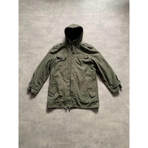 Op de afbeelding: Een olijfgroene jas met capuchon, meerdere zakken en knoopsluitingen. De jas heeft een trekkoord in de taille en manchetten met knopen. De capuchon is gevoerd met zwarte stof. De jas ligt plat op een grijze ondergrond.