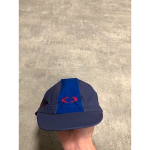 May include: A navy blue cycling cap with a blue center stripe and a red logo. The cap has a curved brim and is made of a lightweight material. The Oakley logo is visible on the front.