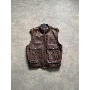 May include: A brown leather vest with multiple pockets and a zipper closure. The vest has a high collar and a quilted lining. It is hanging on a black hanger against a gray background.