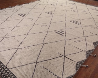 May include: Cream-colored area rug with a geometric diamond pattern. Dark gray lines and triangular shapes create the design. The rug has a dark gray border with tassels on the edge. The rug is on a wooden floor.