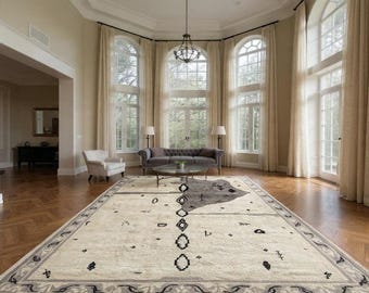 May include: A large, rectangular area rug with a cream-colored field and a gray border. The rug features a central design of black geometric shapes and a gray abstract shape. The room has large windows and a chandelier.