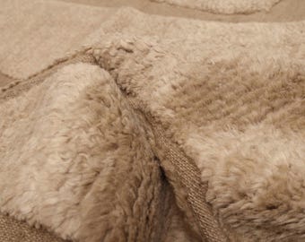 May include: A beige area rug featuring a thick, high-pile shaggy texture contrasted with smooth, flat-woven sections. The design consists of geometric shapes formed by the alternating pile heights, creating a tactile surface with a neutral colour palette.
