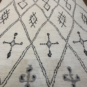 May include: A cream-colored area rug featuring a large black diamond grid pattern. The design includes various gray and black geometric symbols and motifs centered within the diamonds. The rug has a textured surface and short fringe along the edges.