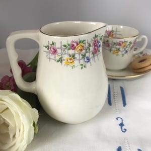 May include: A white ceramic creamer with a floral design featuring pink, yellow, and blue roses. The creamer has a gold rim and a handle.