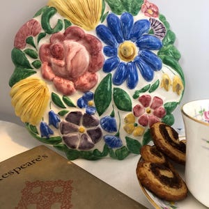 Könnte beinhalten: Eine weiße Keramik-Wandplatte mit einem erhabenen Blumendesign. Die Blumen sind in den Farben Rosa, Blau, Gelb, Grün und Lila bemalt. Die Platte ist blattförmig und hat eine leicht strukturierte Oberfläche.
