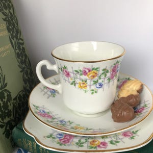Könnte beinhalten: Ein weißes Tee-Set aus Tasse und Untertasse mit einem Blumenmuster aus rosa, gelben und blauen Rosen auf einem Gitter. Das Set hat einen goldenen Rand und ist mit einem zarten Blumenmuster verziert. Das Set steht auf einem grünen Buch.