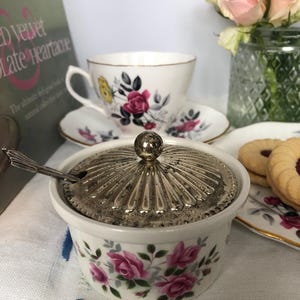 Könnte beinhalten: Eine weiße Zuckerdose mit silbernem Deckel und kleinem Löffel. Die Dose ist mit rosa Rosen und grünen Blättern verziert. Eine Tasse und Untertasse mit passendem Blumendesign befinden sich im Hintergrund, zusammen mit Keksen und Rosen.