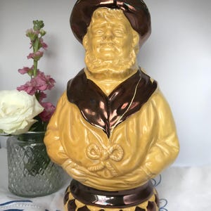 Könnte beinhalten: Vintage Keramikfigur eines Mannes mit Bart, der einen gelben Mantel und einen braunen Hut trägt. Die Figur hat eine glänzende Oberfläche und ist mit braunen Akzenten verziert. Der Sockel ist ebenfalls braun mit dekorativen Elementen.