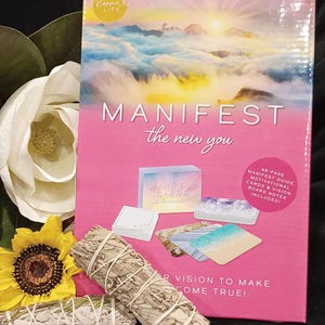 以下が含まれることがあります： ピンクと白の箱に「MANIFEST the new you」という文字と、雲の上の夕焼けの画像が描かれています。箱の中には、マニフェステーションカードとガイド、ビジョンボードのメモが入っています。