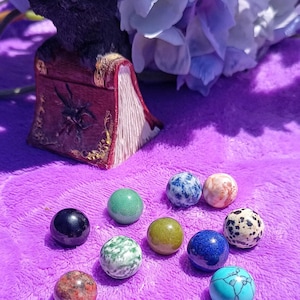 May include: A collection of 11 colourful, smooth, round, polished stones on a purple surface. The stones are various shades of green, blue, red, pink, and white.