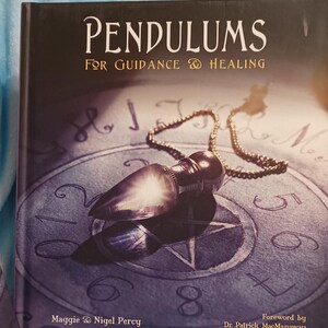 May include: A book titled "Pendulums for Guidance & Healing" with a silver pendulum on a chain resting on a purple and white background with numbers and symbols. The book has a foreword by Dr. Patrick MacManaway.