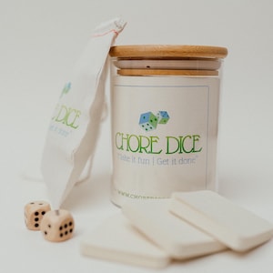 Op de afbeelding: Een glazen pot met een houten deksel met dobbelstenen erin. De pot heeft een wit etiket met de tekst "CHORE DICE" en de slogan "Make it fun | Get it done". De pot staat op een wit oppervlak met een witte stoffen zak, dobbelstenen en witte kaarten.