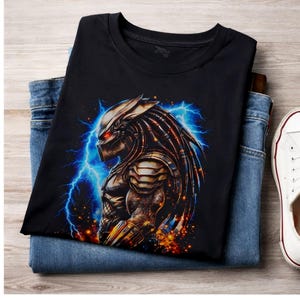 Könnte beinhalten: Schwarzes T-Shirt mit einer detaillierten Illustration eines Predator-Charakters mit roten Augen, vor einem Hintergrund aus blauem Blitz. Das T-Shirt ist auf einer blauen Jeans gefaltet, ein weißer Sneaker ist sichtbar.
