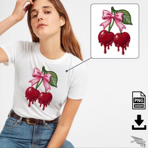 Puede incluir: Camiseta blanca con un diseño bordado de dos cerezas rojas, lazos rosas y hojas verdes. Las cerezas tienen un efecto goteo. El diseño está en la parte delantera de la camiseta. La modelo lleva vaqueros azules.