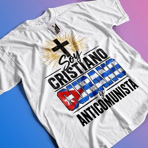 Puede incluir: Camiseta blanca con un diseño gráfico que presenta una cruz negra, un sol y texto en español: "Soy Cristiano Cubano y Anticomunista". La bandera cubana está incorporada en el diseño. La camiseta está sobre un fondo degradado azul y rosa.