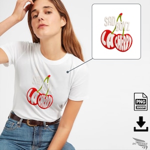 Puede incluir: Camiseta blanca con un diseño de cereza roja y el texto "SAD BOYZ LA CHERRY". Las cerezas tienen tallos verdes y el texto está en una fuente blanca estilizada. La camiseta se combina con vaqueros azules y un cinturón marrón.