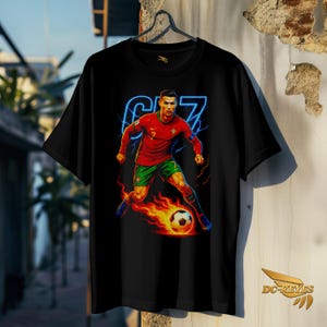May include: Black t-shirt featuring a vibrant graphic of a soccer player in a red jersey with green shorts, kicking a flaming soccer ball. The design includes the number 7 and the player's name in blue neon. The shirt is hanging on a wall.