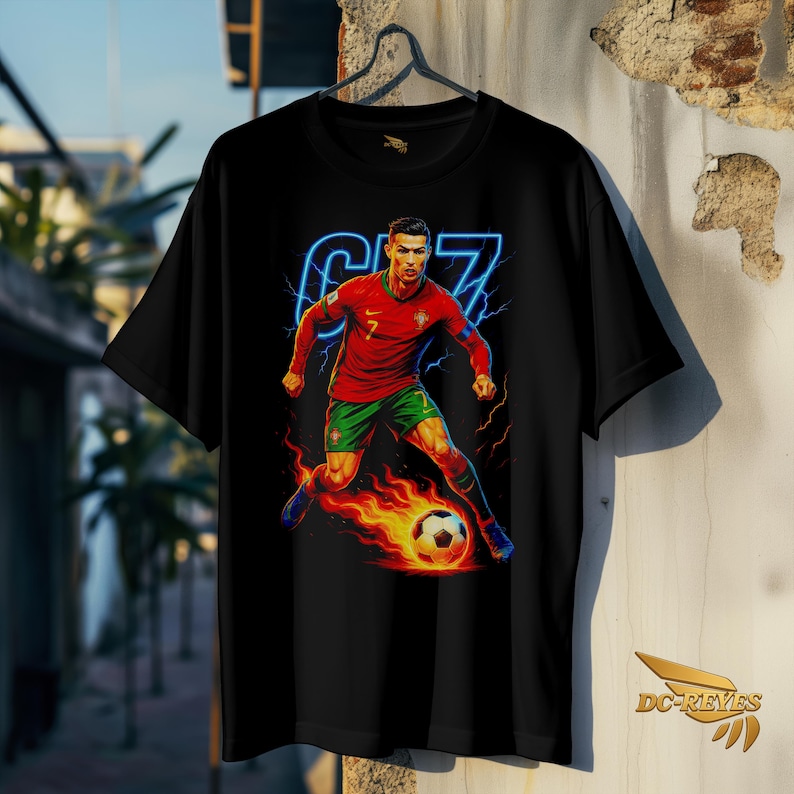 May include: Black t-shirt featuring a vibrant graphic of a soccer player in a red jersey with green shorts, kicking a flaming soccer ball. The design includes the number 7 and the player's name in blue neon. The shirt is hanging on a wall.