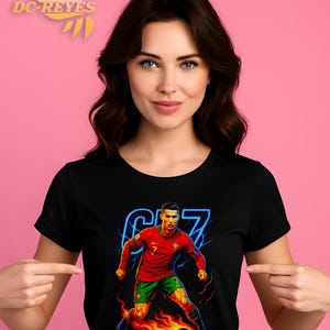 May include: Black t-shirt featuring a vibrant graphic of a soccer player in a red and green uniform, with the number 7 and the player's image. The design includes a soccer ball engulfed in flames and a neon blue outline. The shirt is worn by a person.
