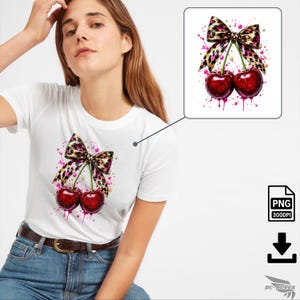 Puede incluir: Camiseta blanca con un gráfico de dos cerezas y un lazo con estampado de leopardo. Las cerezas son de un rojo intenso con salpicaduras de pintura rosa. El lazo tiene un diseño de estampado de leopardo rosa y marrón.