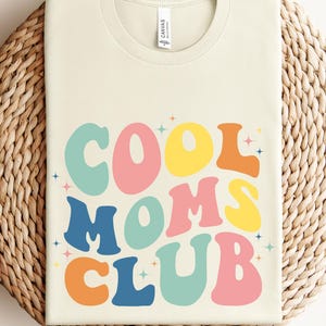 Puede incluir: Una camiseta beige con el texto "COOL MOMS CLUB" en letras coloridas de estilo retro. Las letras están delineadas en blanco y tienen un ligero efecto de sombra.