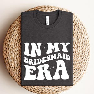 May include: A dark gray t-shirt with the text "In My Bridesmaid Era" in white, retro-style lettering with sparkling stars.
