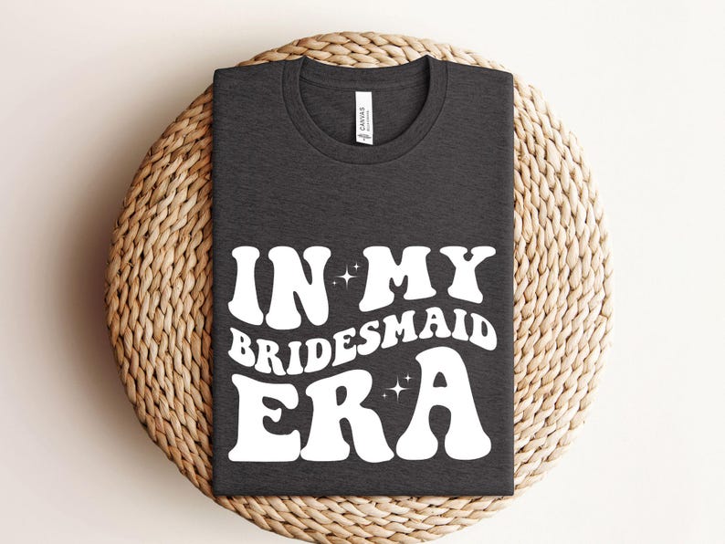 May include: A dark gray t-shirt with the text "In My Bridesmaid Era" in white, retro-style lettering with sparkling stars.