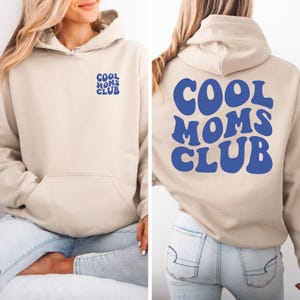 Puede incluir: Una sudadera con capucha beige con un gráfico azul "Cool Moms Club" en la espalda y una versión más pequeña del gráfico en el bolsillo delantero.