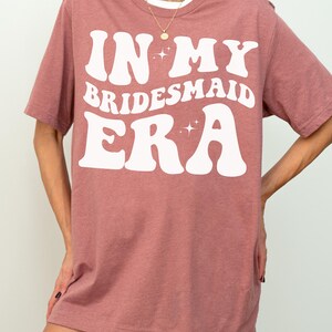 May include: A heather mauve t-shirt with the text "In My Bridesmaid Era" in white retro-style lettering with sparkling stars.