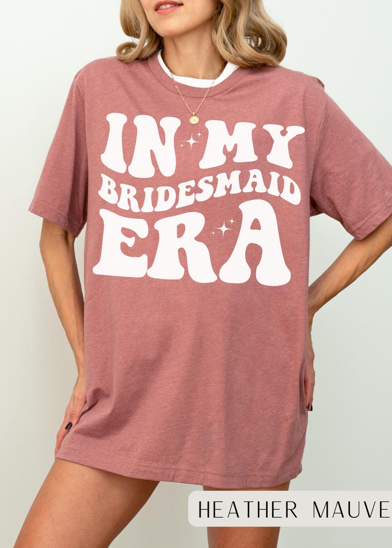 May include: A heather mauve t-shirt with the text "In My Bridesmaid Era" in white retro-style lettering with sparkling stars.