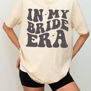 May include: A soft cream colored t-shirt with the text "IN MY BRIDE ERA" in black, retro-style lettering. The text is surrounded by small, sparkly stars.