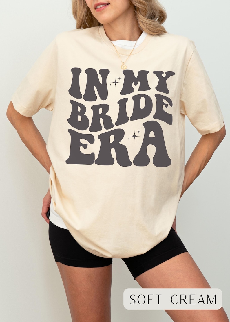 May include: A soft cream colored t-shirt with the text "IN MY BRIDE ERA" in black, retro-style lettering. The text is surrounded by small, sparkly stars.
