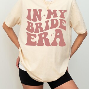 May include: A cream-colored t-shirt with the text "IN MY BRIDE ERA" in pink retro-style lettering. The shirt is oversized and has short sleeves.