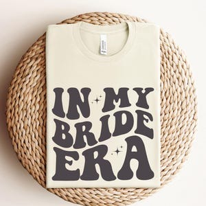 May include: A white t-shirt with the text "In My Bride Era" in black, retro-style lettering. The text is surrounded by small, sparkly stars.