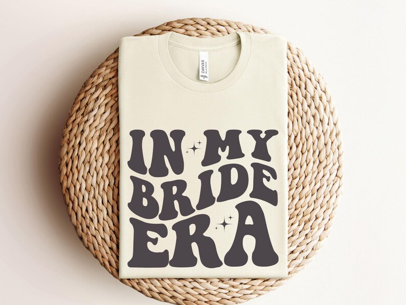 May include: A white t-shirt with the text "In My Bride Era" in black, retro-style lettering. The text is surrounded by small, sparkly stars.