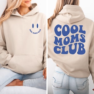 Puede incluir: Una sudadera con capucha beige con una carita sonriente azul en la parte delantera y el texto "Cool Moms Club" en azul retro en la parte trasera.