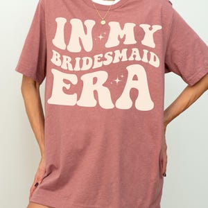 May include: A heather mauve t-shirt with the text "In My Bridesmaid Era" in a retro font with sparkling stars.